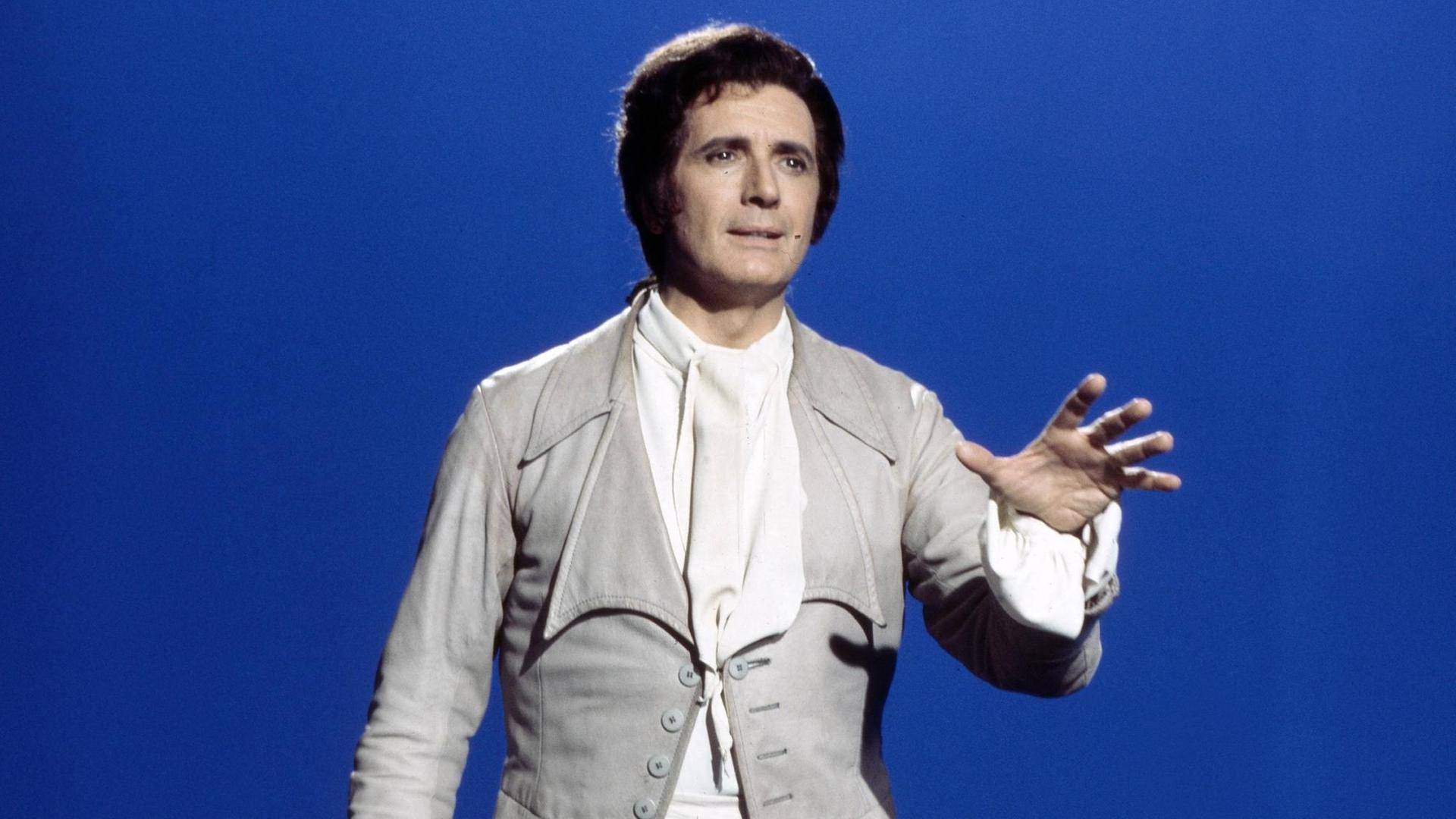 Der Sänger Franco Corelli steht vor einem mittelblauen Hintergrund, er trägt ein Kostüm im Stil des 18. Jahrhunderts, der linke Arm ist nach vorne ausgestreckt.