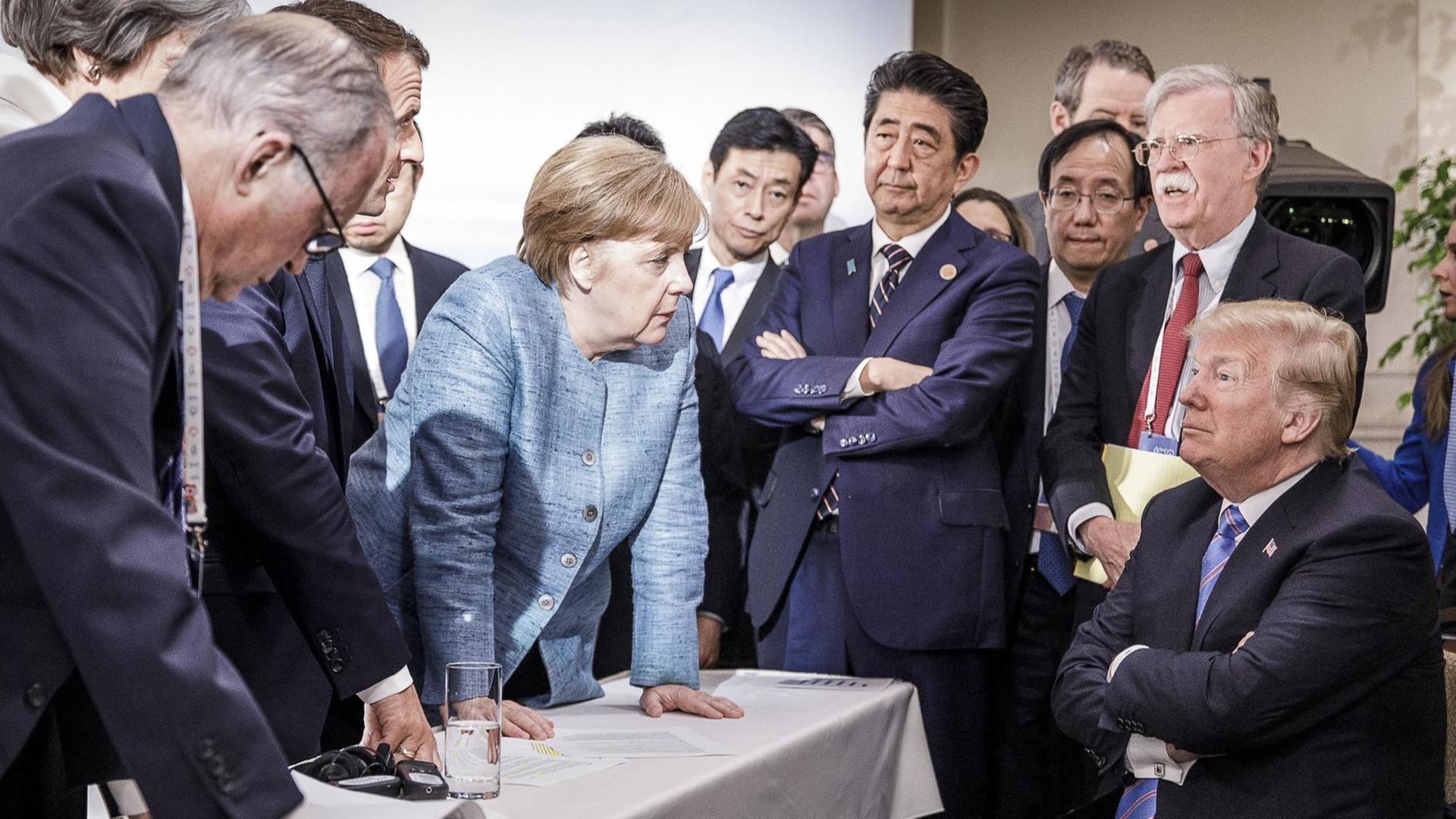 Angela Merkel und Donald Trump während des G7-Gipfels. Merkel steht vor Trump, aufgestützt auf einen Tisch. Trump sitzt als Einziger und schaut an Merkel vorbei
