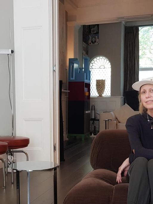 Die Kunstsammlerin Valeria Napoleone sitzt auf einem Sessel und blickt in die Kamera. Links von ihr ist ein Gemälde oder ein bearbeitetes Foto an der Wand. Darauf ist eine junge Frau zu sehen, die auf einem Bürostuhl sitzt. Ihre Strumpfhose ist bis zu den Knien heruntergezogen.