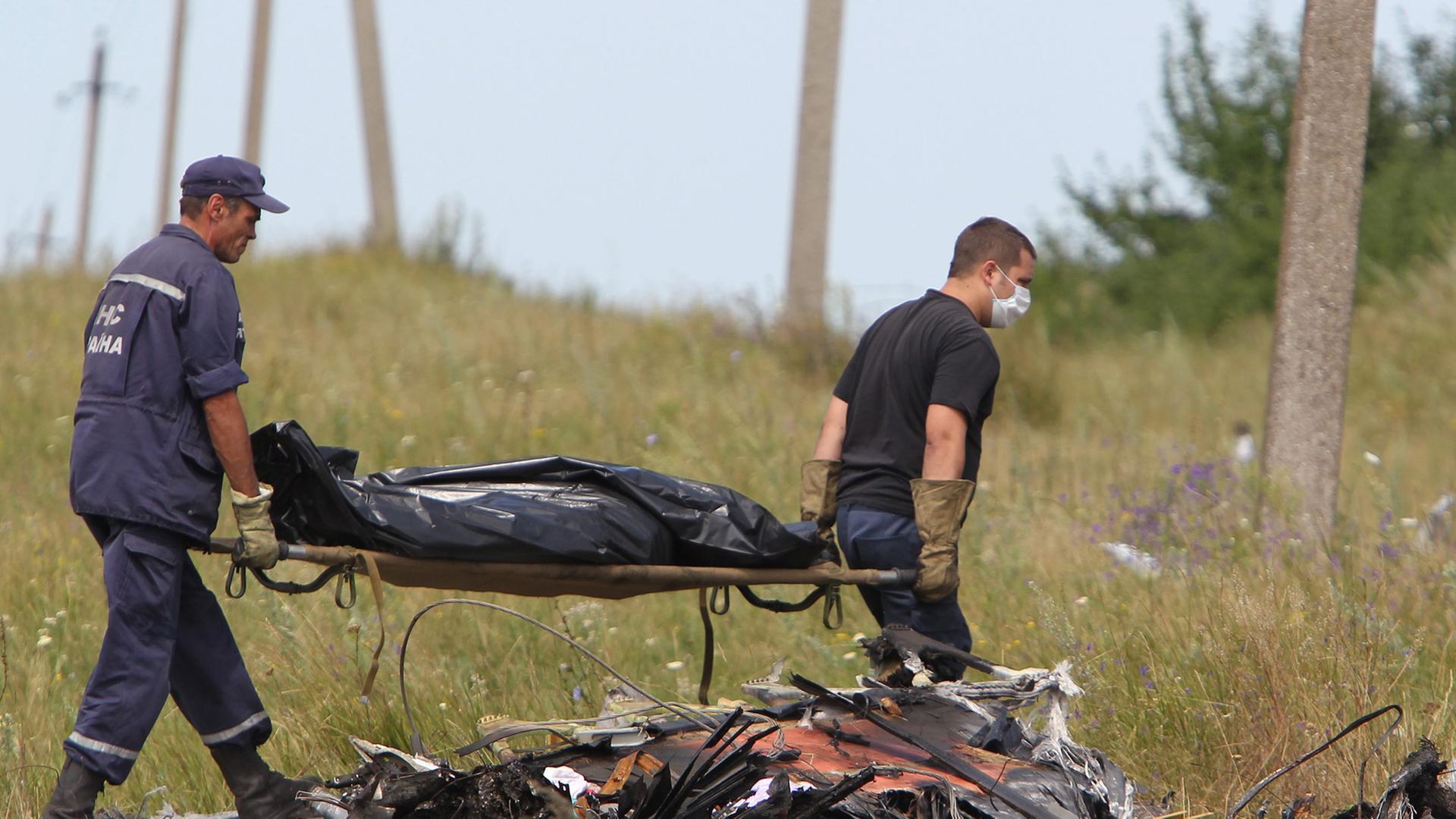 Hinter Trümmern tragen zwei ukrainische Arbeiter eine Bahre, auf der ein abgedecktes Todesopfer liegt.