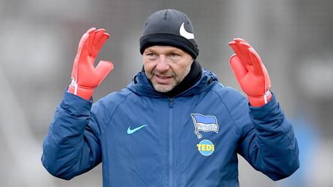 Torwarttrainer Zsolt Petrysteht mit Mütze, Torwarthandschuhen und Herta-Jacke auf dem Platz und hebt die Hände neben den Kopf.