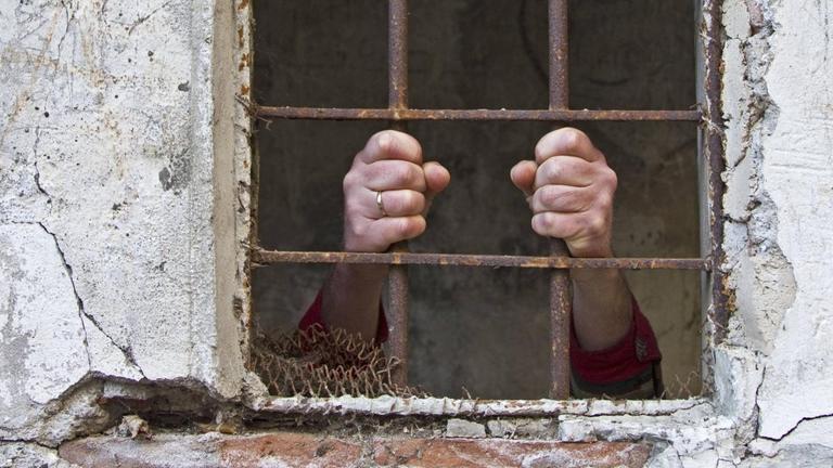 Die Hände eines Gefangenen an einem Gitterfenster.
