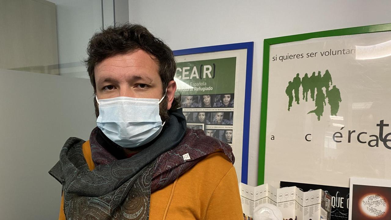 Ein junger Mann mit dunklem Haar, gelbem Pulli  und Mundschutz sowie einem Halstuch steht in seinem Büro.