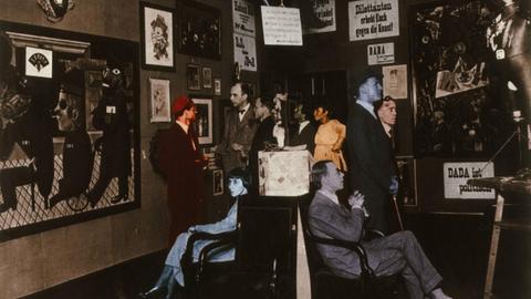 Eröffnung der 1. Internationalen Dada-Messe, Berlin, Buchhandlung Dr. Burchard, 5. Juli 1920 (v. li. stehend Raoul Hausmann, Otto Burchard, Baader, Wieland und Margarete Herzfelde, George Grosz, John Heartfield; sitzend Hannah Höch und Otto Schmalhausen. Foto, spätere Kolorierung.