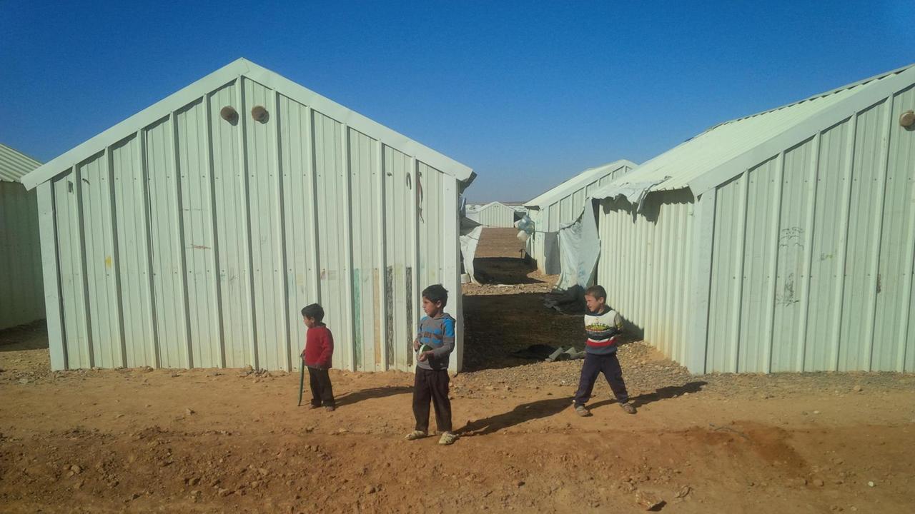 Drei kleine Jungen stehen vor hellgrünen Wellblechhütten im Flüchtlingslager Azraq in Jordanien.