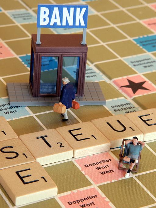 Eine Miniaturfigur trägt zwei Geldkoffer auf einem Scrabble-Spiel in eine Bank (Illustration), auf dem das Wort Steueroase liegt.