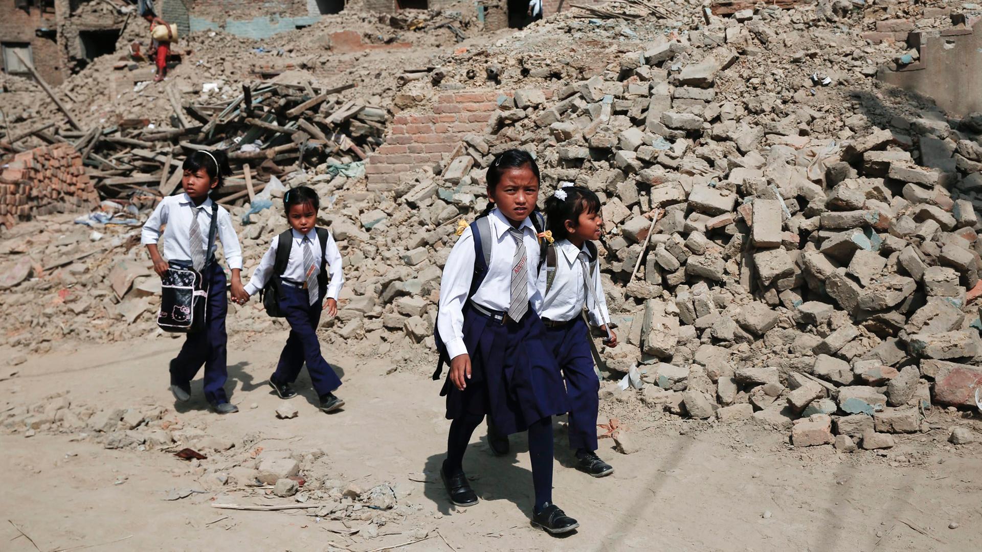 Kinder in Nepal auf dem Weg zur Schule. Die Kinder tragen eine Schul-Uniform. Sie laufen an einem zerstörten Haus vorbei.