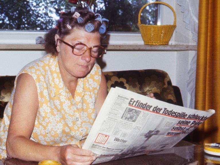 Eine Frau mit Lockenwicklern im Haar und in Kittelschürze sitzt Ende der 70er-Jahre auf einer Couch und liest eine Zeitung.