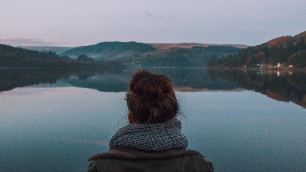 Panoramablick über einen ruhigen Bergsee. Eine Frau blickt auf den See, sie steht mittig im Bild und mit dem Rücken zur Kamera.