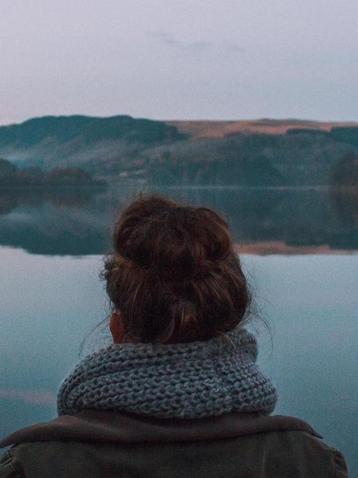 Panoramablick über einen ruhigen Bergsee. Eine Frau blickt auf den See, sie steht mittig im Bild und mit dem Rücken zur Kamera.
