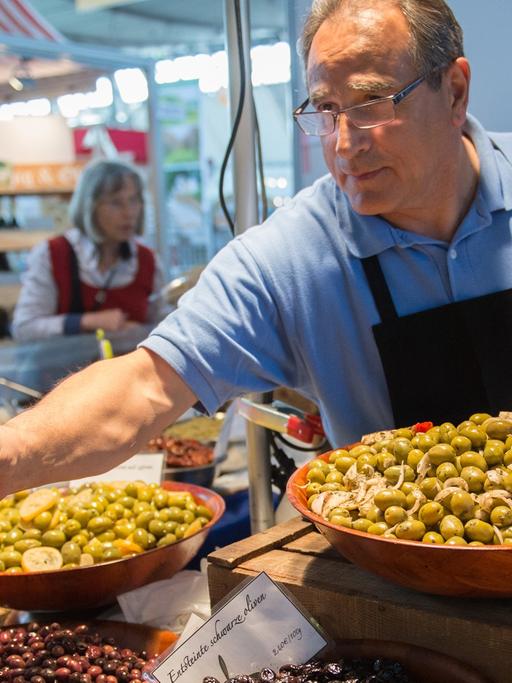 Ein Mann mit Schürze steht hinter einem Verkaufsstand mit Oliven und füllt welche davon in eine Tüte.