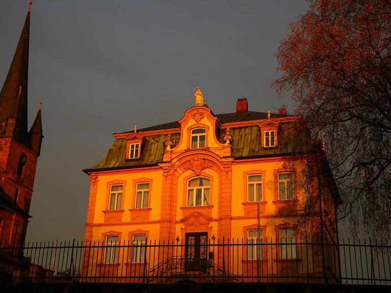 Die untergehende Sonne lässt die Kirche St. Wenzeslaus in Bayern und das zugehörige Pfarrhaus, einen neubarocken Bau im Villenstil orange aufleuchten.