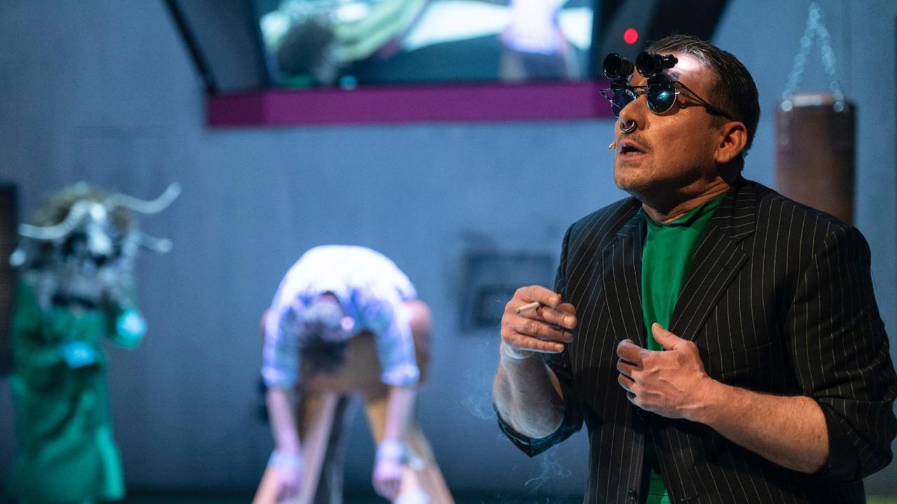 Bühnenszenario: Im Vordergrund steht ein Schauspieler mit Zigarette in der Hand. Er trägt Sonnenbrille mit einem aufgesteckten Fernglas. Im Hintergrund in der Unschärfe weitere Schauspieler.
