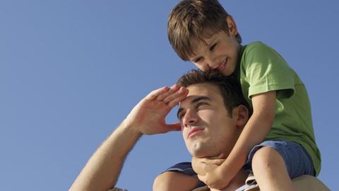 Ein Junge sitzt auf den Schultern seines Vaters, der seine Hand an der Stirn hat und in die Ferne blickt.
