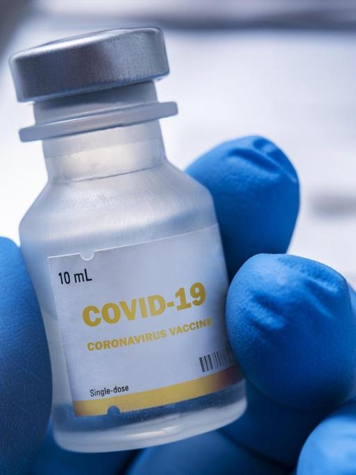 Nahaufnahme einer behandschuhten Hand, die ein Fläschchen mit der Aufschrift "Covid-19 - Coronavirus Vaccine" hält