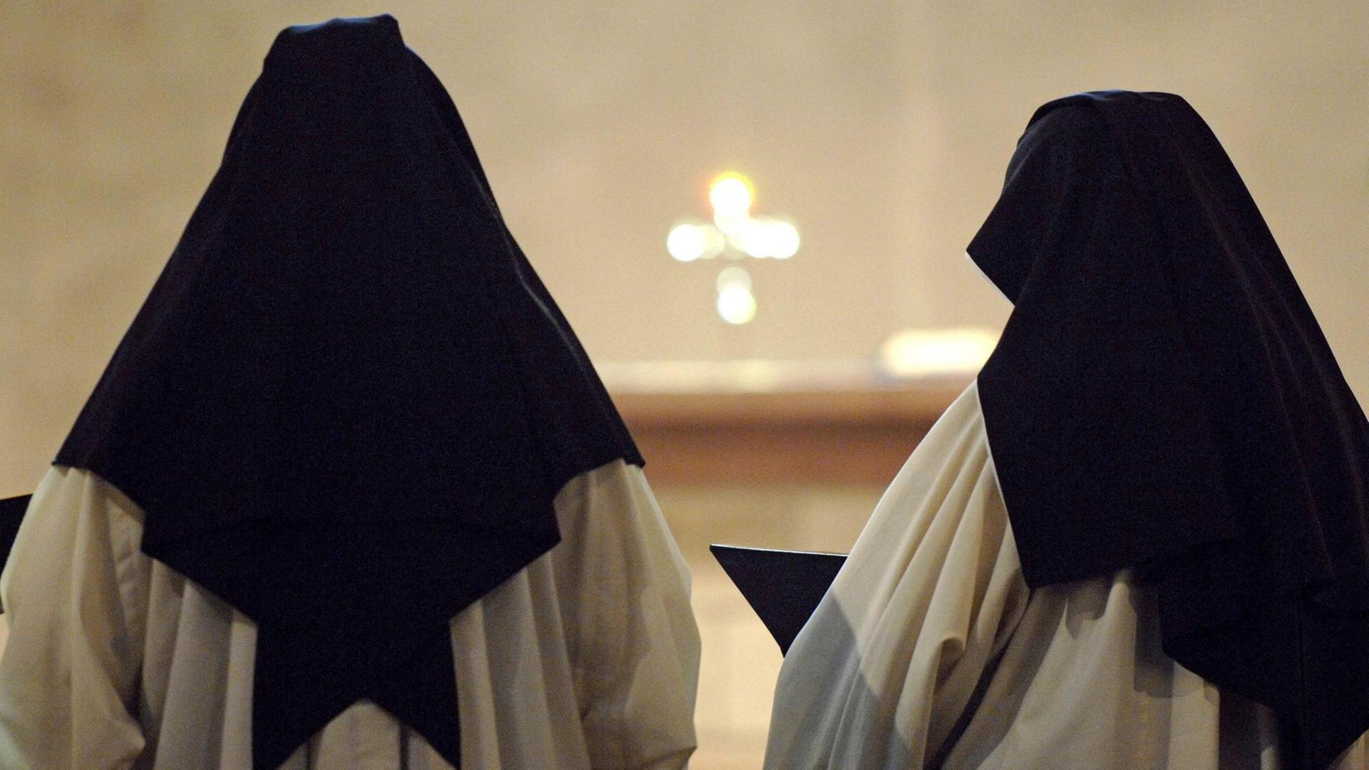 Zwei Zisterzienserinnen stehen ein einem Kloster in Deutschland bei der Messe mit dem Rücken zur Kamera. Sie tragen ein weißes Gewand und einen schwarzen Nonnenschleier. 