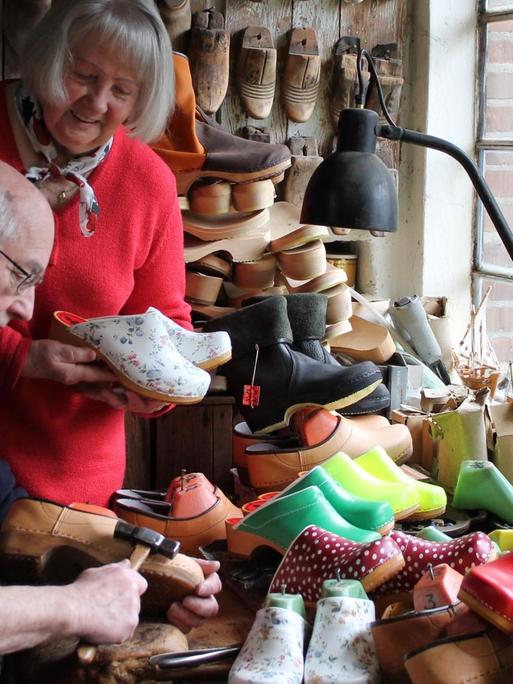 Blick in die Werkstatt von Schuster Lorenz Hamann, der an einem Tisch voller Holzschuhe sitzt und einen Schuh bearbeitet, hinter ihm steht eine Frau mit rotem Pullover und hält ein Paar Holzschuhe in der Hand