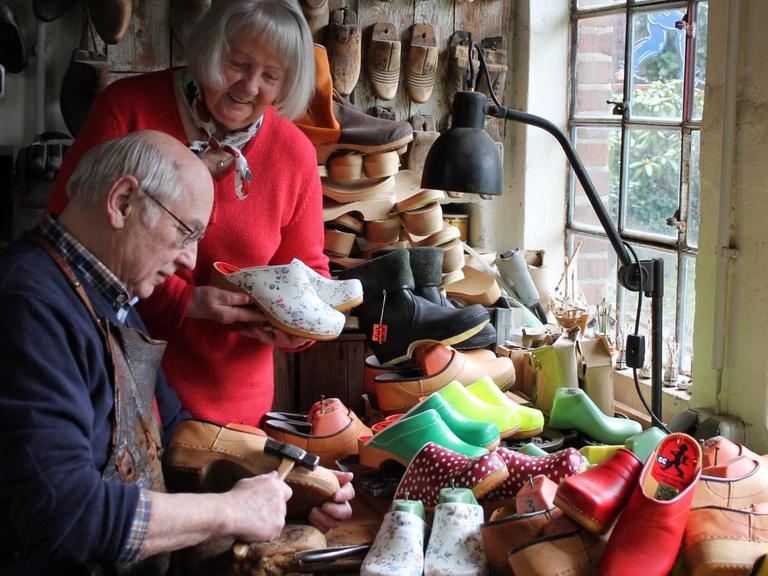 Blick in die Werkstatt von Schuster Lorenz Hamann, der an einem Tisch voller Holzschuhe sitzt und einen Schuh bearbeitet, hinter ihm steht eine Frau mit rotem Pullover und hält ein Paar Holzschuhe in der Hand