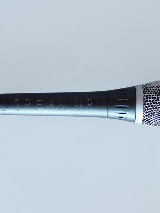 Ein Mikrofon vor grauem Hintergrund. Darauf eingeritz steht "Speak Up".