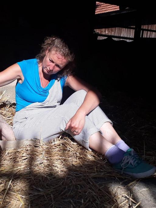 Eine blonde Frau sitzt in einem Stall auf Stroh. Neben ihr liegt ein Schwein.