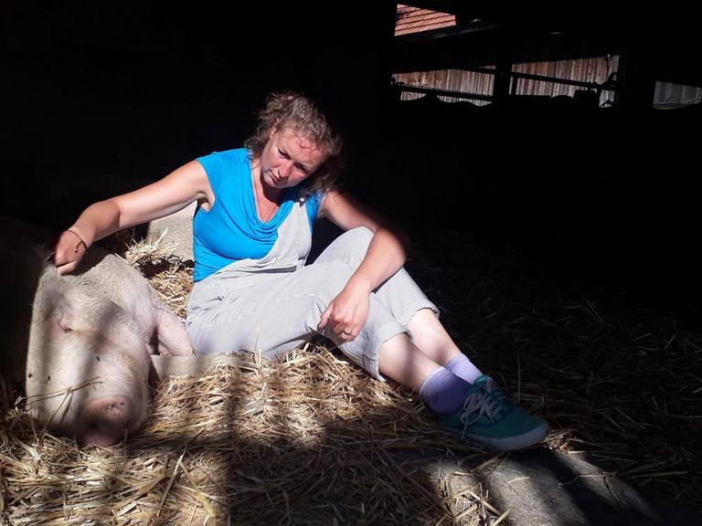 Eine blonde Frau sitzt in einem Stall auf Stroh. Neben ihr liegt ein Schwein.