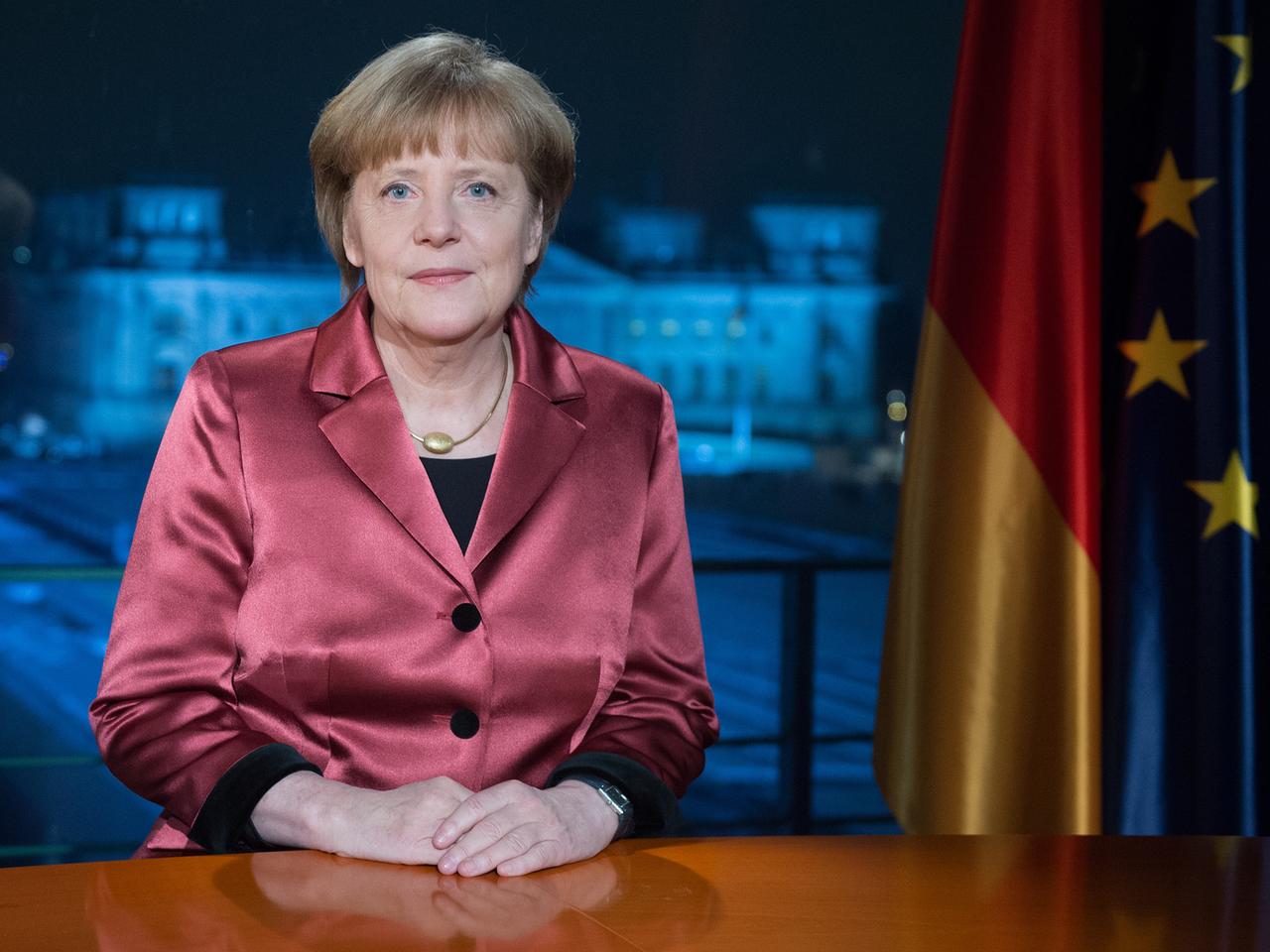 Angela Merkel an einem Schreibtisch vor Deutschland- und EU-Flagge, im Hintergrund der Blick durchs Fenster auf den Reichstag.