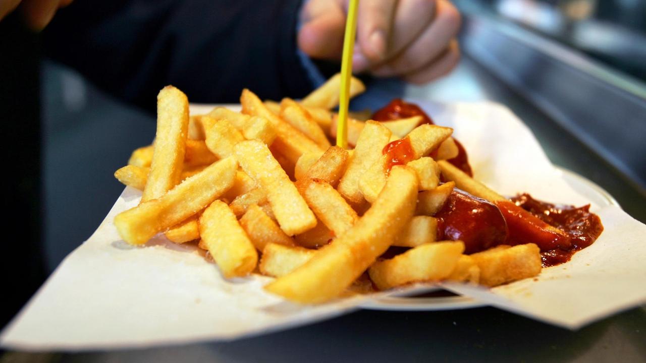 Auf einem Tisch liegt eine eine Portion Pommes Frittes mit Ketchup. Eine Hand sticht gerade mit einer Pommesgabel in eine Fritte.