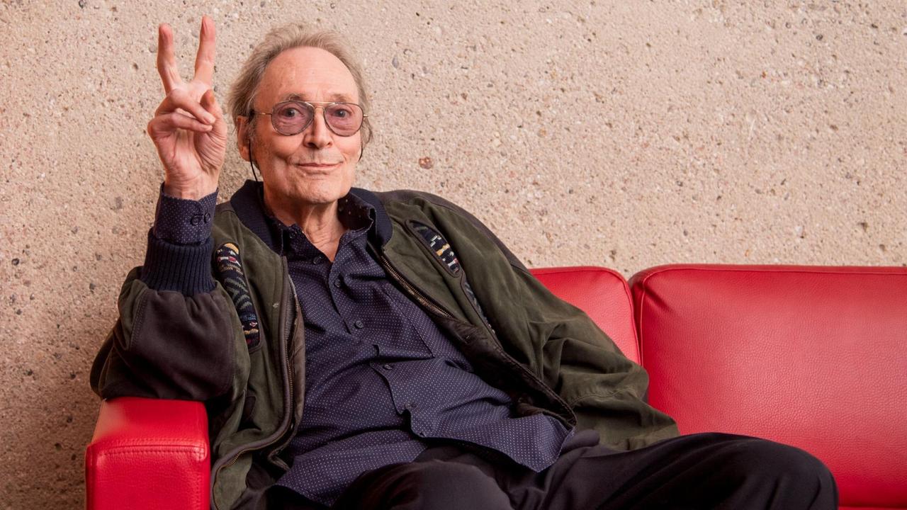 Regisseur Roland Klick, ein älterer Mann mit Brille, sitzt auf einem roten Sofa und zeigt das Peace-Zeichen in die Kamera.