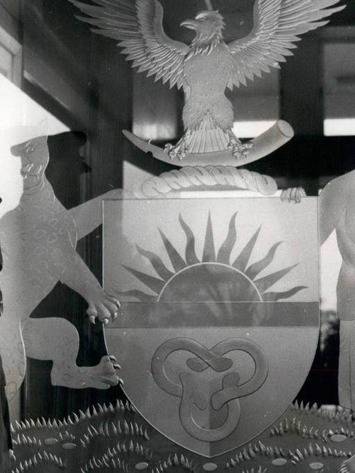 Ein historisches Schwarz-Weiß-Foto. Zu sehen ist links ein afrikanischer Mann mit Vollbart und in Militäruniform. Er steht vor dem Symbol des damaligen Staates Biafra im Südosten von Nigeria. Das Symbol zeigt unter anderem einen Adler und eine aufgehende Sonne.