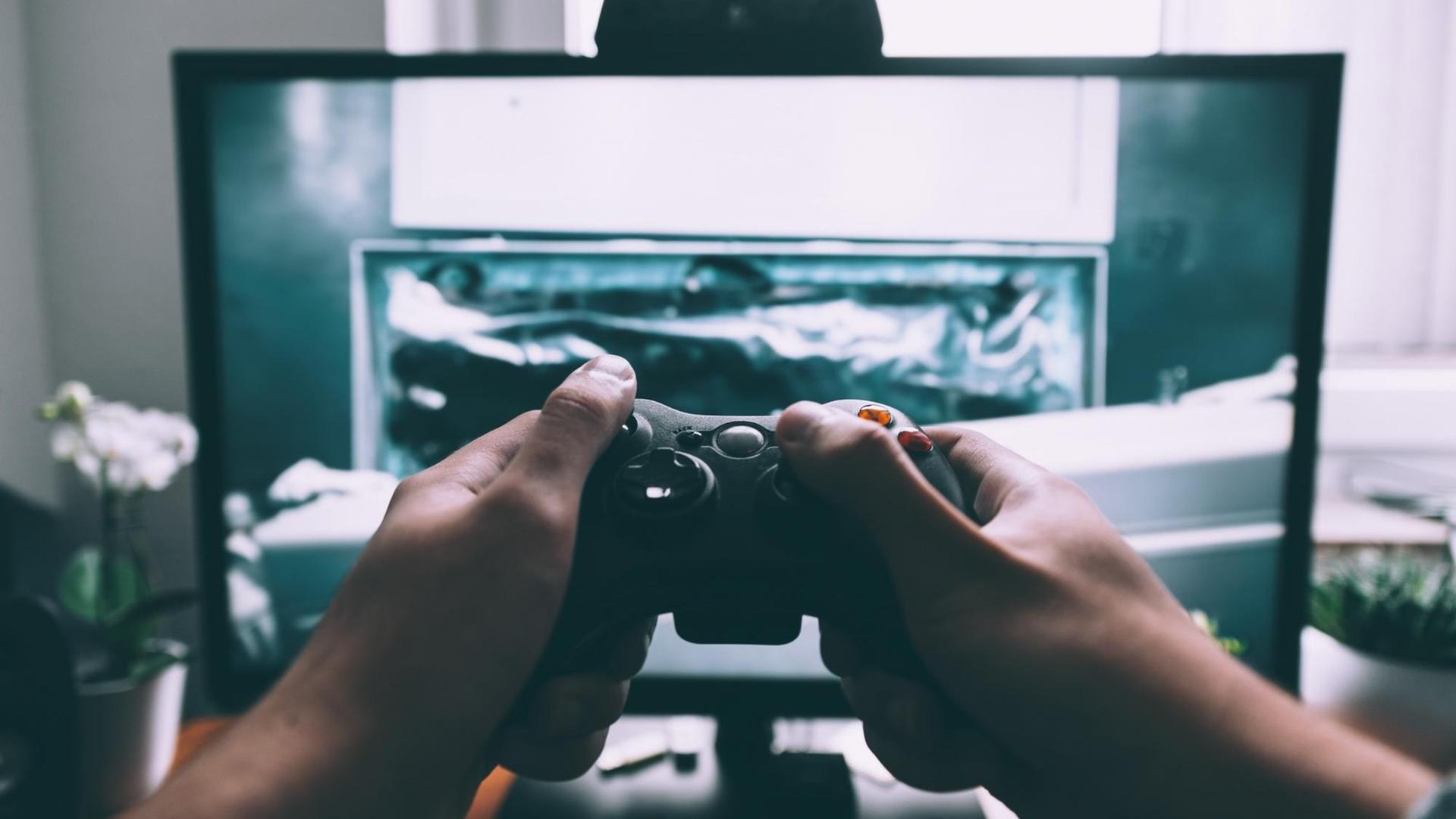 Ein Fernseher, auf dem ein Computerspiel läuft, im Vordergrund Hände, die eine Spielekonsole halten.