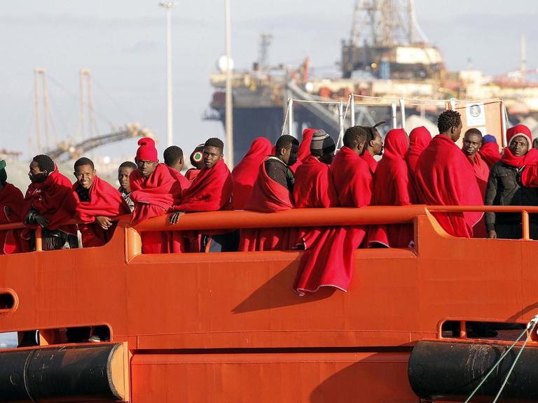 Gerettete Flüchtlinge aus der Subsahara stehen auf einem Boot der spanischen Küstenwache, eingewickelt in rote Decken.