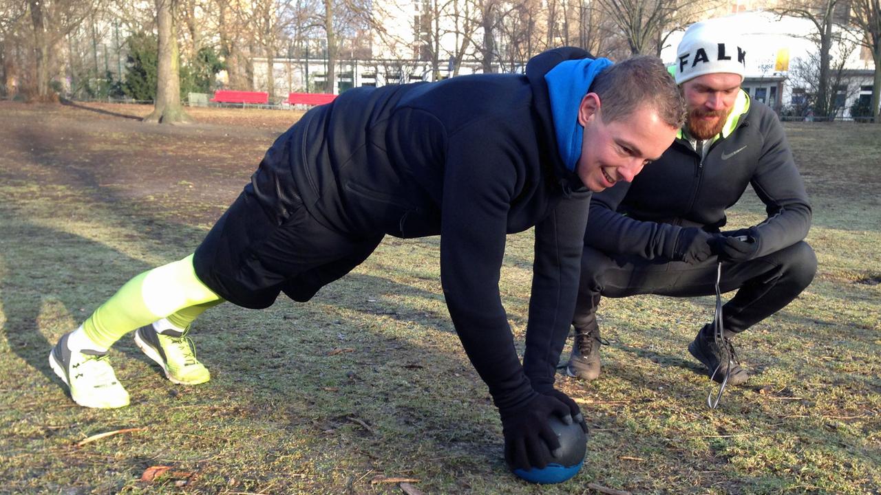 Ein Mann in Trainingskleidung absolviert auf einen Ball gestützt Fitnessübungen im Park. Neben ihm hockt sein Trainer Michel Gleich in Laufkleidung und mit einer Stoppuhr.
