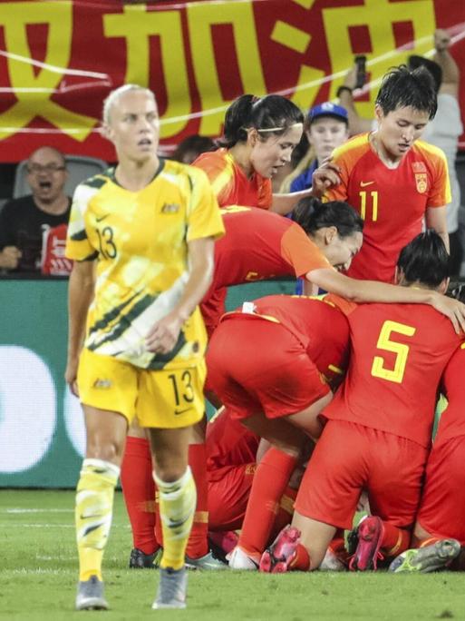 Die chinesische Fußballolympiamannschaft feiert ein Tor gegen Australien. Im Hintergrund chinesische Fans mit einem Transparent in Rot mit gelber Schrift.
