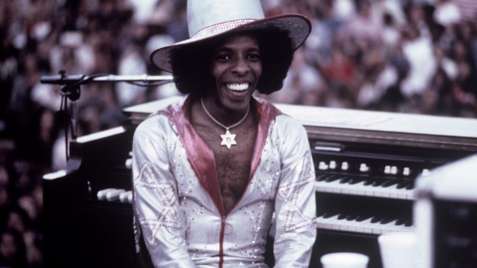 Der US-Musiker Sly Stone sitzt 1974 in einem weißglänzenden Anzug bei einem Auftritt auf einer Bühne.