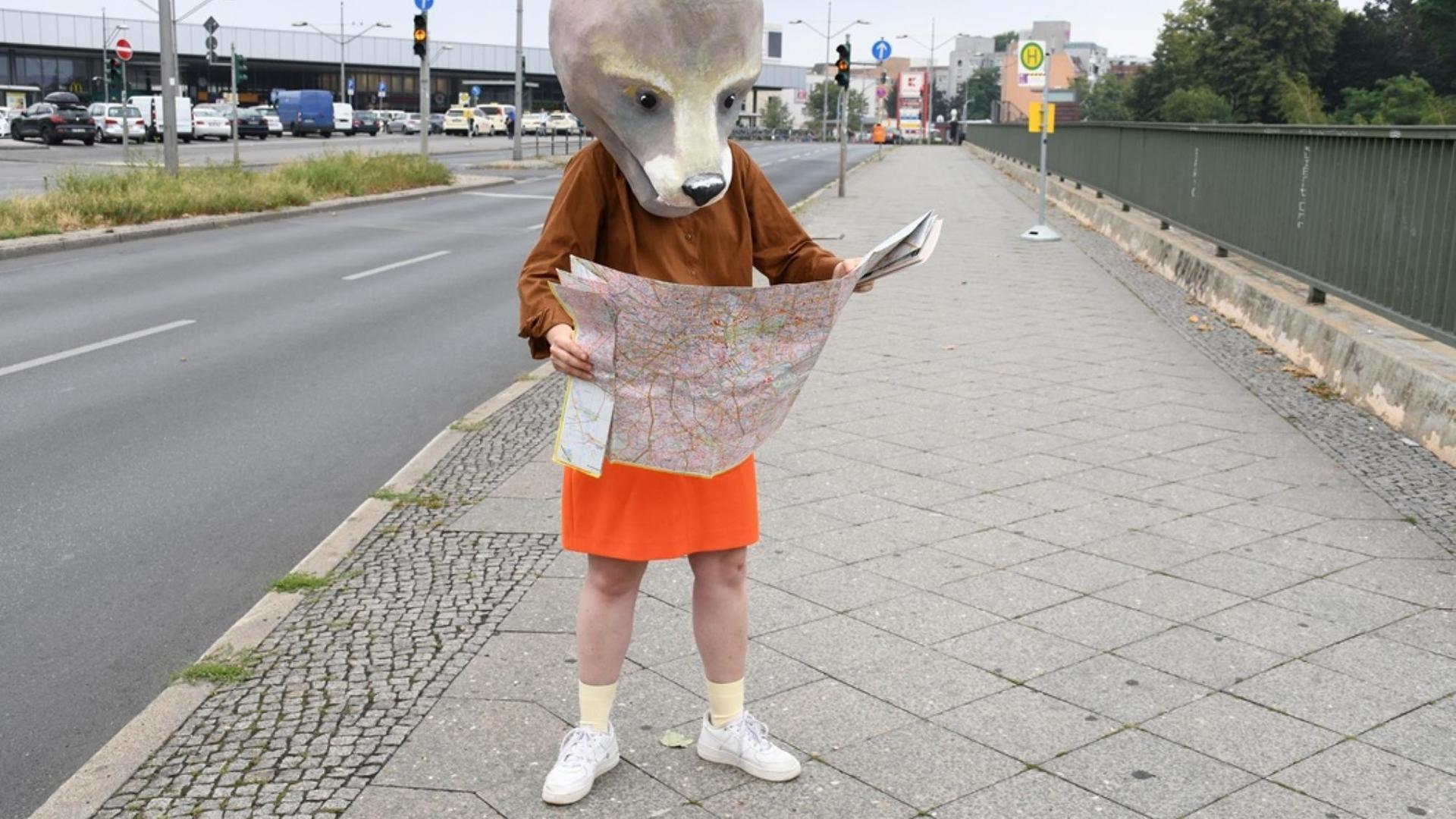 Ein Mensch mit einem Bärenkopf steht auf der Straße und hält einen Stadtplan fest.