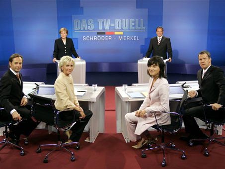 Die beiden Kontrahenten Angela Merkel und Gerhard Schröder stehen in einem Studio in Berlin Adlershof. Im Vordergrund: die Moderatoren Thomas Kausch, Sabine Christiansen, Maybrit Illner und Peter Klöppel.