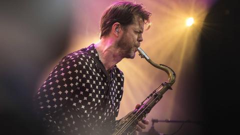 Im Gegenlicht eines Bühnenscheinwerfers spielt ein bärtiger Mann mittleren Alters Tenorsaxofon.
