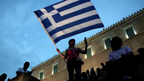 Eine Frau steht während der Demonstration auf einer Mauer vor dem Abendhimmel. Sie hält eine griechische Flagge.