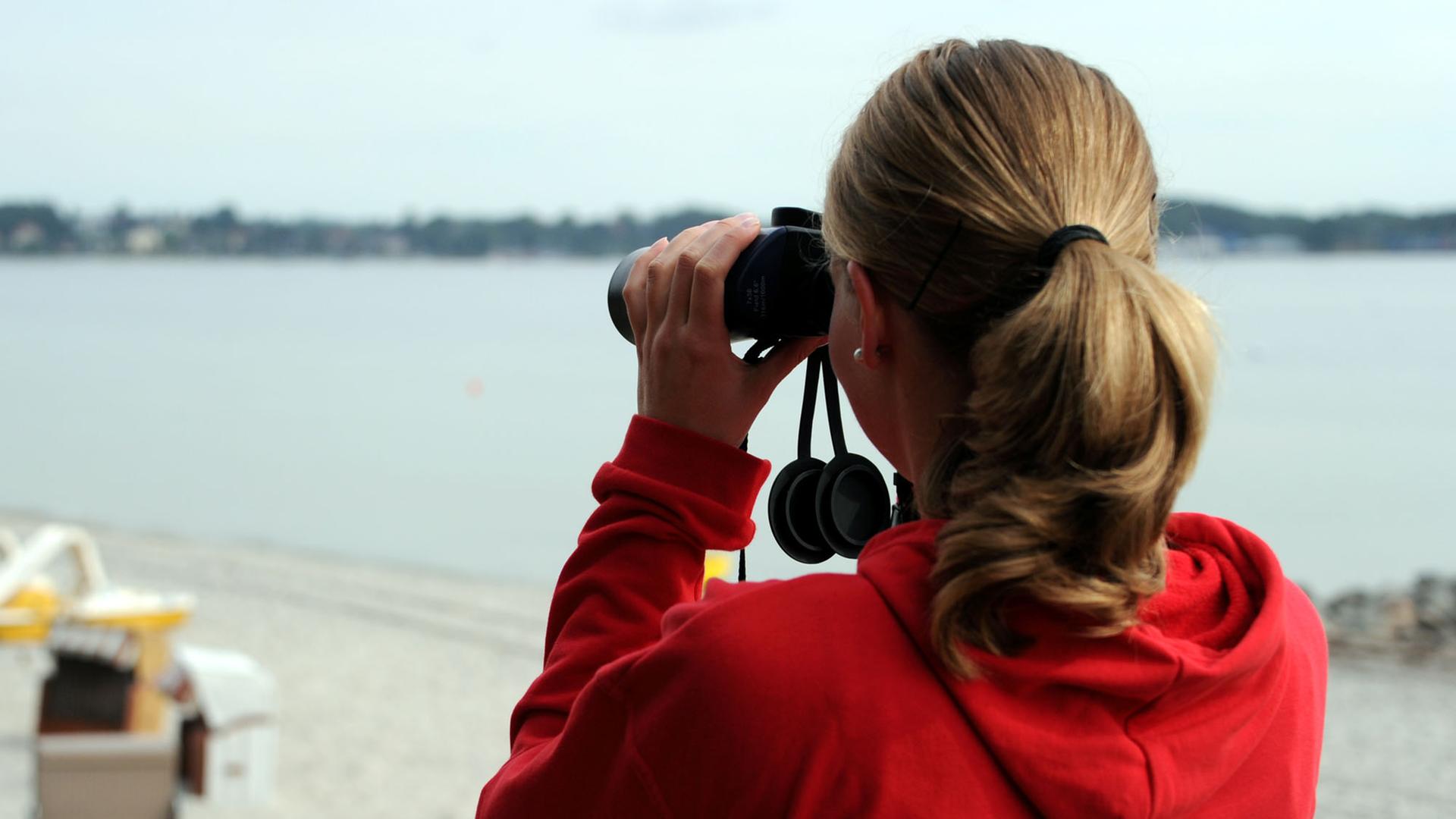 Ein freiwilliges Mitglied der Deutschen Lebensrettungsgesellschaft (DLRG) blickt am 19.08.2013 mit einem Fernglas von der Station auf den Strand von Eckernförde (Schleswig-Holstein). Ein freiwilliges Mitglied der Deutschen Lebensrettungsgesellschaft (DLRG) blickt am 19.08.2013 mit einem Fernglas von der Station auf den Strand von Eckernförde (Schleswig-Holstein).