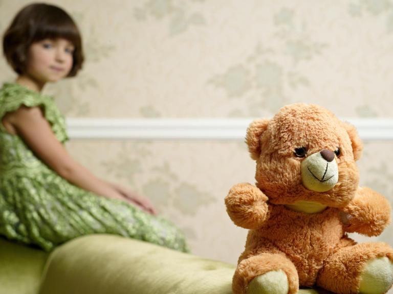 Ein alter Teddy sitzt auf einer Sofalehne, im Hintergrund ist ein Mädchen zu sehen.
