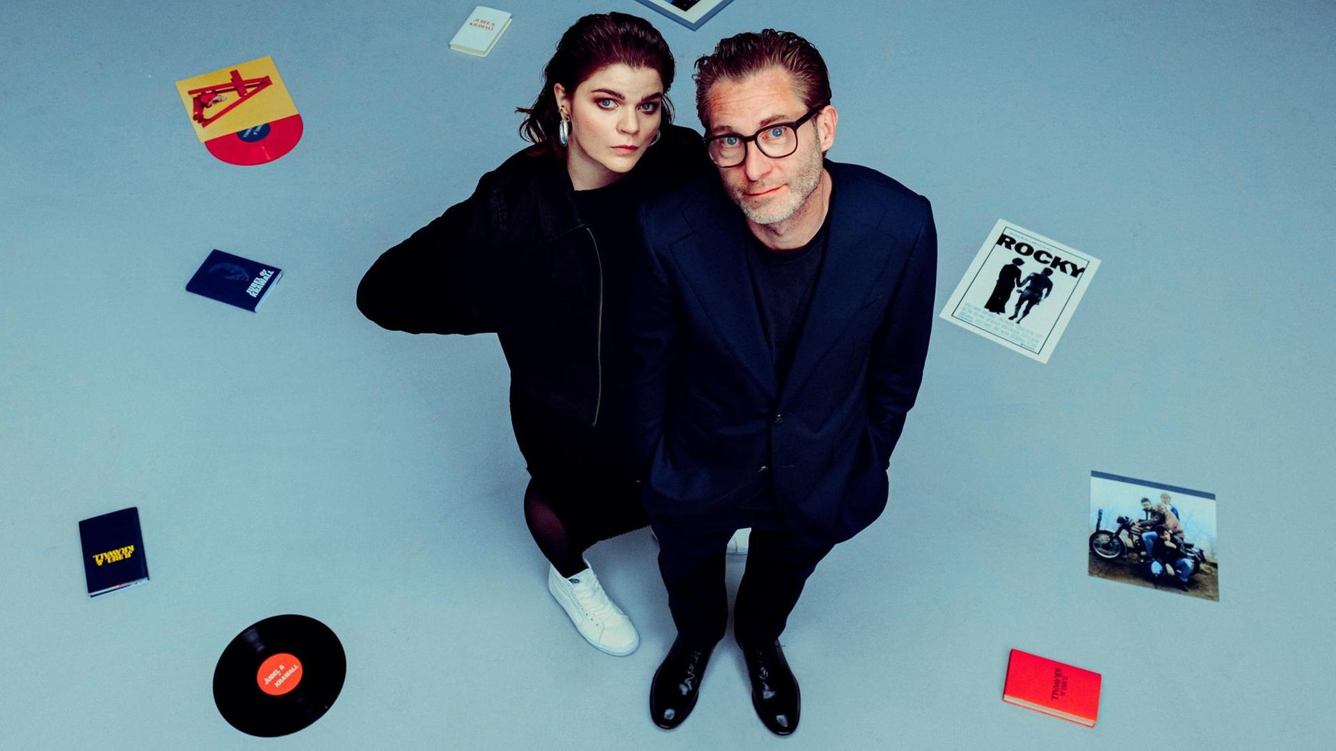 Sophie Passmann und Matthias Kalle stehen in dunkler Kleidung inmitten von Schallplatten, CDs und Büchern.