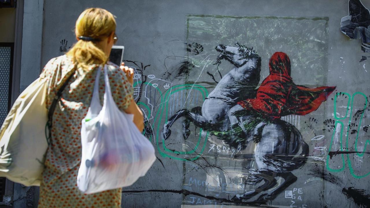 Streetart des britischen Künstlers Bansky in Paris und eine Frau, die das Kunstwerk fotografiert.

