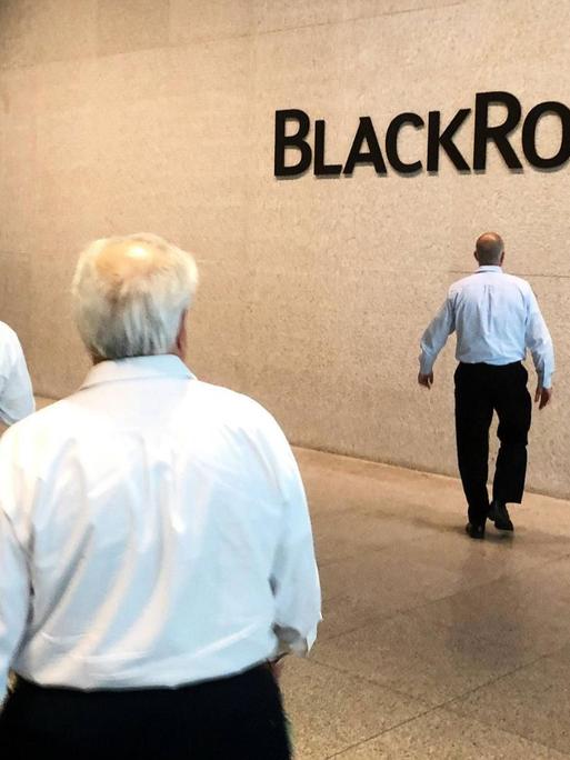 Firmensitz von BlackRock in New York. Drei Männer laufen in einer Halle, im Hintergrund steht an einer Wand BlackRock.