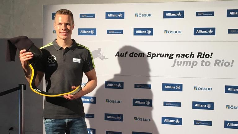 Der Weitspringer Markus Rehm hält eine Bein-Spring-Prothese und lächelt in die Kamera, im Hintergrund eine Wand, auf der steht: Auf dem Sprung nach Rio.