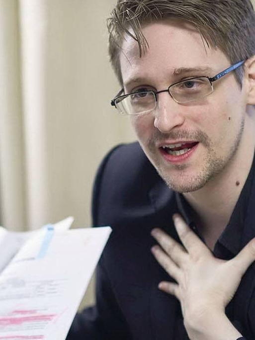 Snowden sitzt auf einem Sessel und zeigt ein Dokument