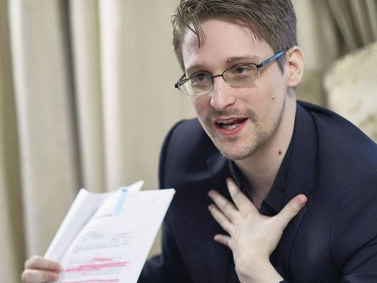 Snowden sitzt auf einem Sessel und zeigt ein Dokument