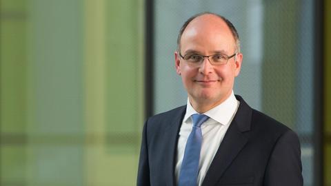 Stefan Gerwens steht im Anzug vor einem grafischen Hintergrund und blickt in die Kamera.