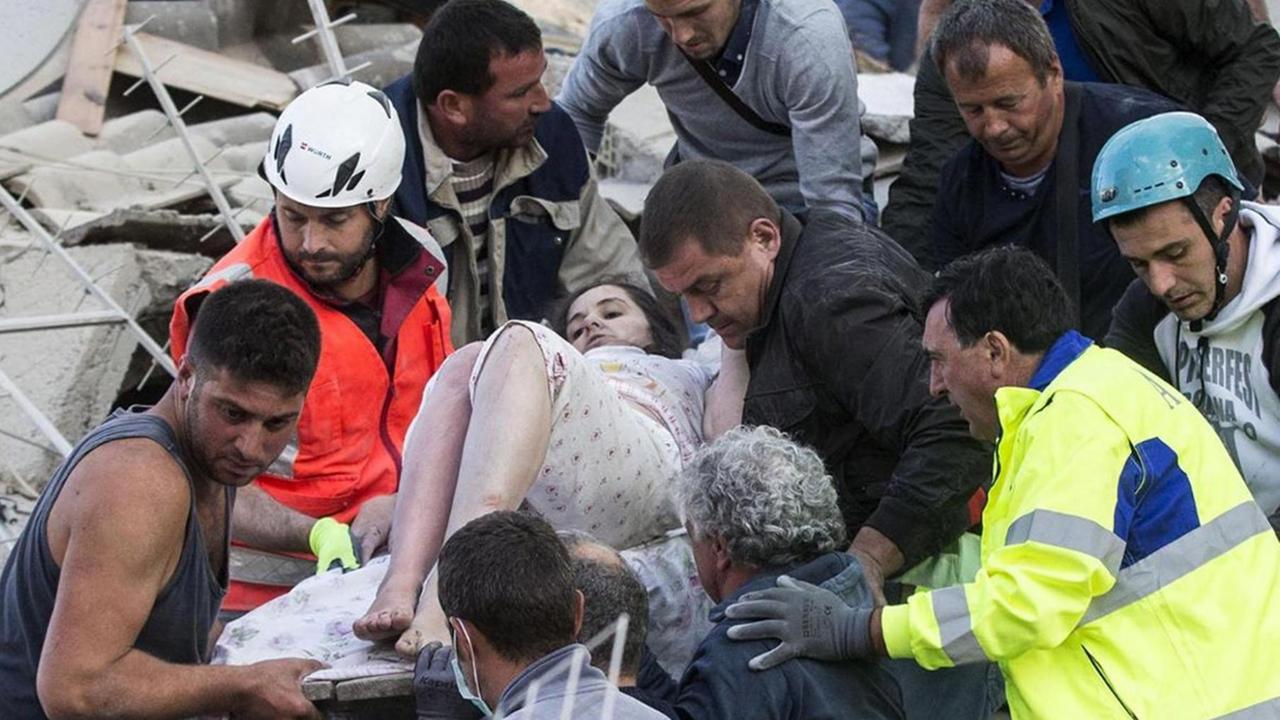 Die staubbedeckte Frau wird von zahlreichen Männern auf einer Bahre von einem Trümmerhaufen heruntergetragen.