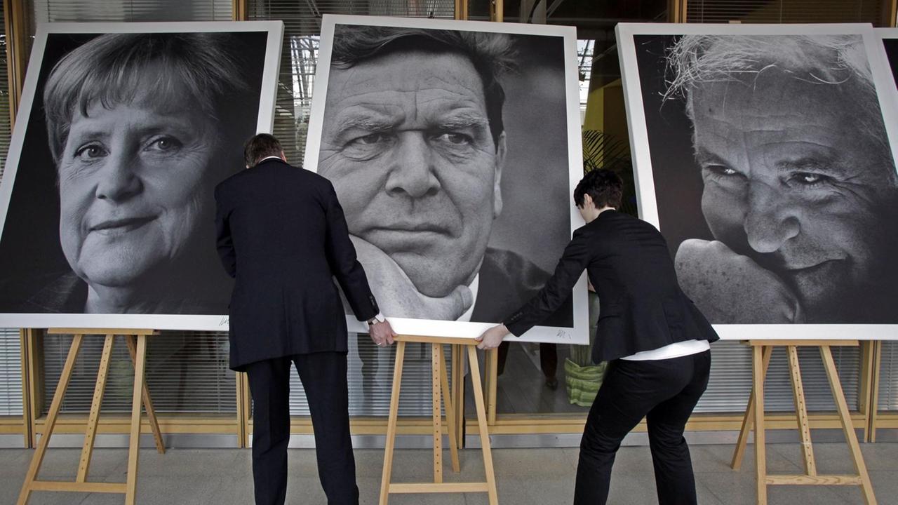 Die Portraits von Angela Merkel, Gerhard Schröder und Helmut Kohl werden aufgerichtet.