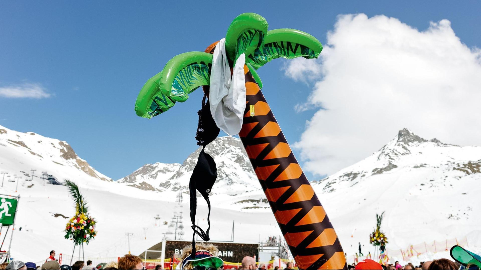 Eine aufblasbare Palme, an der ein BH hängt, steht vor einem schneebedecktem Alpenpanorama.